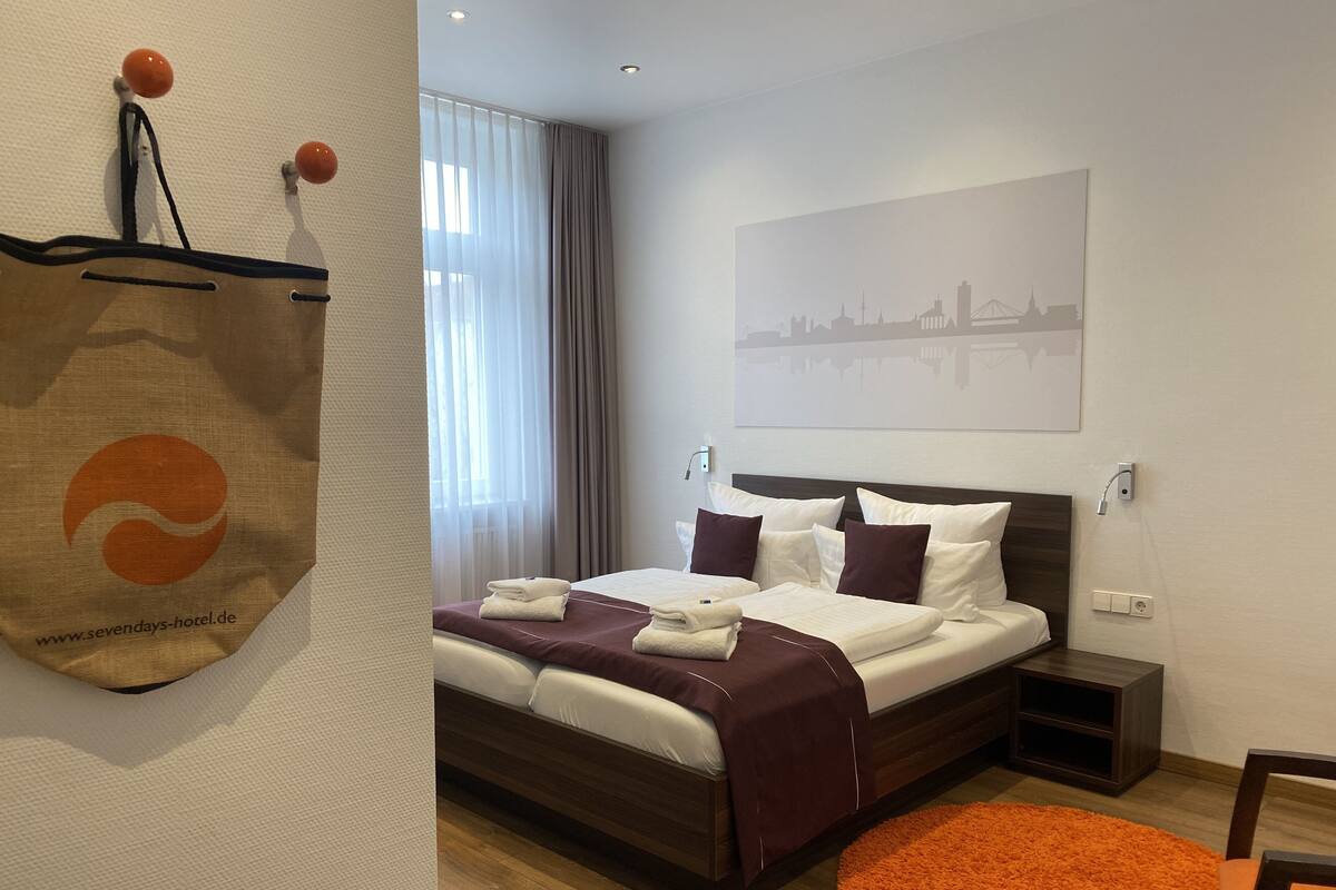 Habitación de hotel con cama, mesita de noche, una pequeña mesa, una silla, ventana con cortinas y un cuadro de un paisaje urbano en la pared.