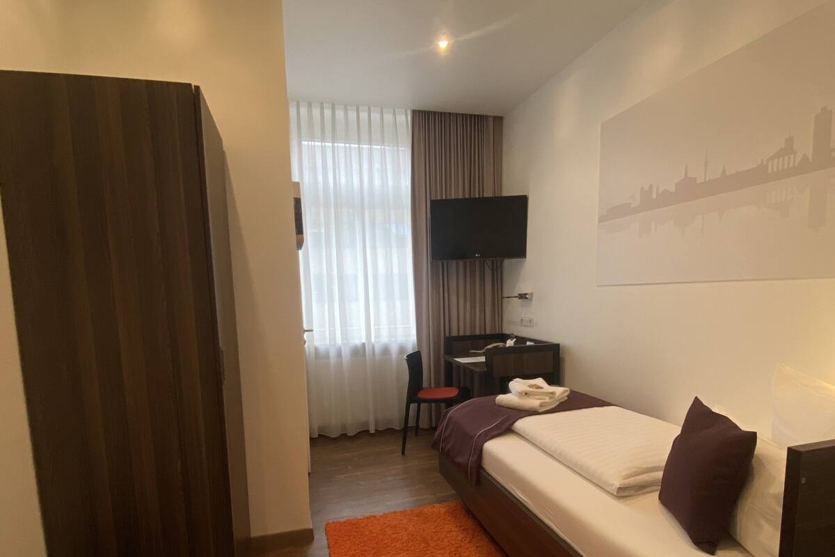 Habitación de hotel con cama, escritorio, silla, televisor y un cuadro de paisaje urbano en la pared.