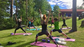 Un groupe de personnes pratiquant le yoga sur l’herbe, avec des arbres et un bâtiment en arrière-plan.