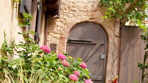 Un bâtiment en pierre avec une porte en bois, entouré d’un chemin de gravier et de plantes en fleurs.