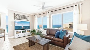 A living room with a TV, a brown leather sofa, a wooden coffee table, and a view of the ocean through large windows. - TIDEWATER 4th FLOOR - SKIP ELEVATORS! No Windows on Hallway, walk to Pier Park! (Panama City Beach)