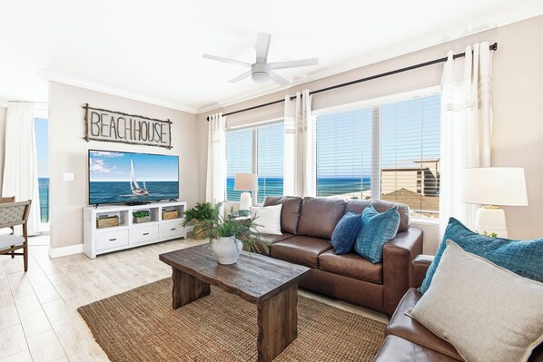 A living room with a TV, a brown leather sofa, a wooden coffee table, and a view of the ocean through large windows.