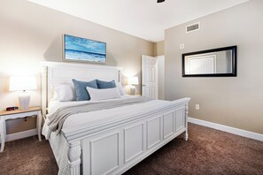 A bedroom with a white bed, beige walls, a painting of a beach, and a door.