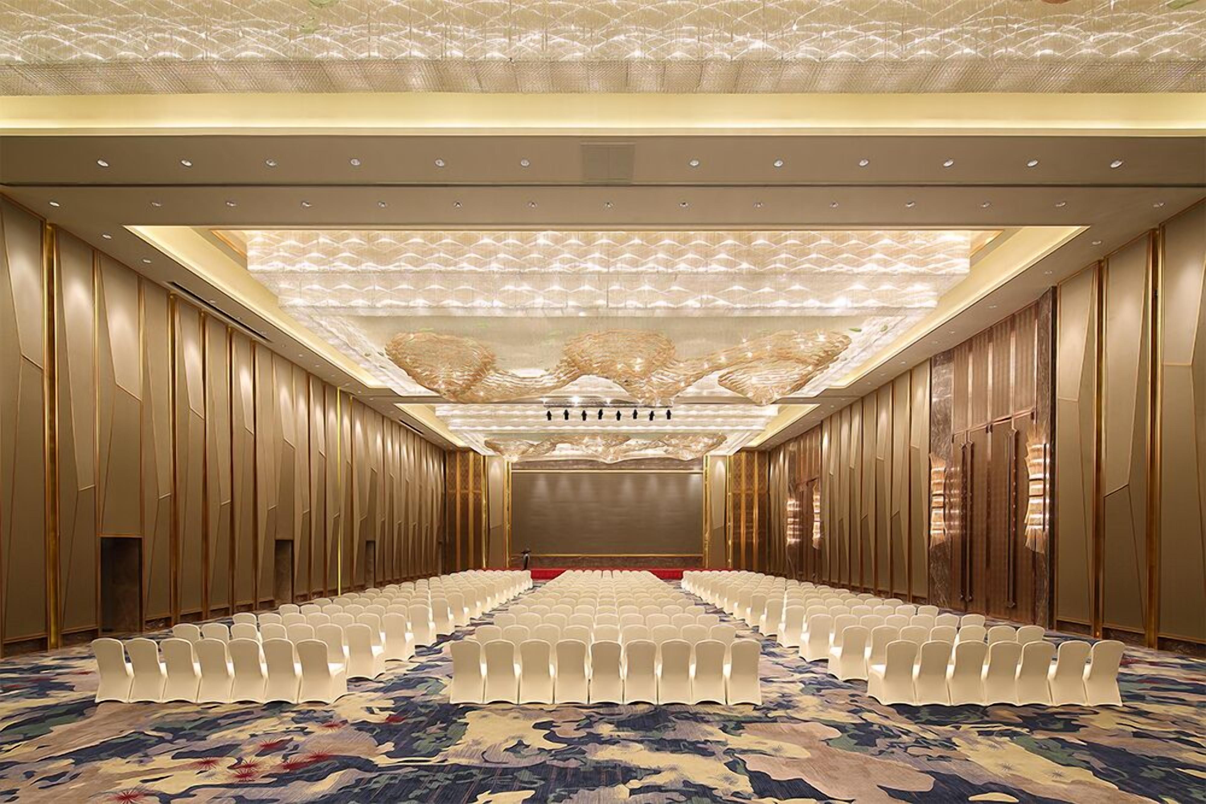 Un salón de conferencias con filas de sillas blancas, una alfombra estampada y un techo decorativo.