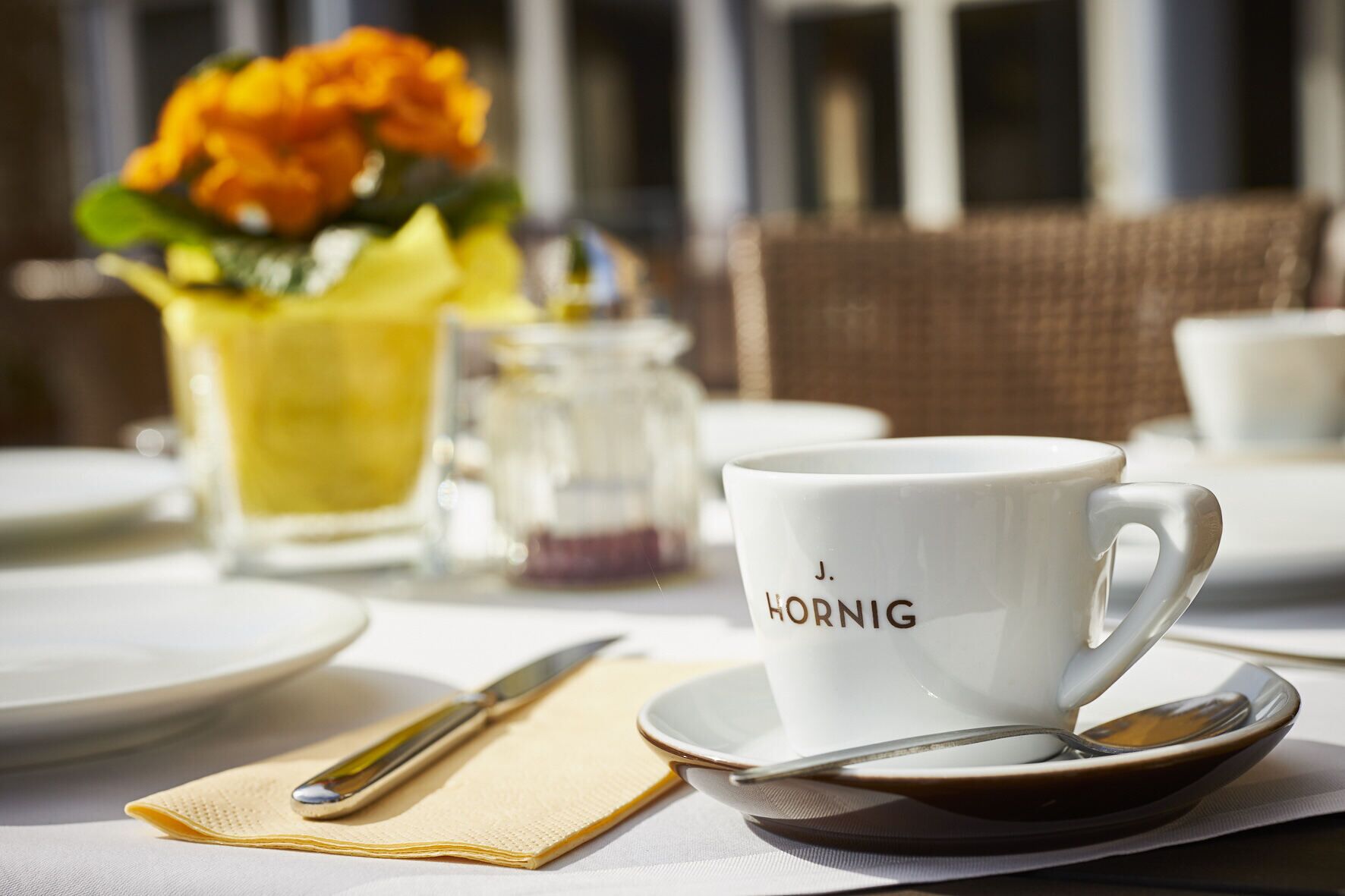 Una mesa dispuesta con una taza blanca que dice "J. HORNIG", una jarrón de vidrio, un florero y una silla de mimbre.