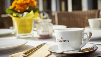 Una mesa dispuesta con una taza blanca que dice "J. HORNIG", una jarrón de vidrio, un florero y una silla de mimbre.
