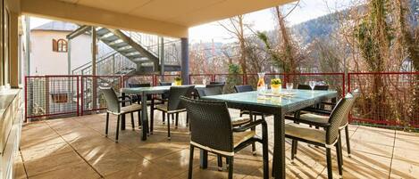 An outdoor patio with a glass table and wicker chairs, a red railing, and a view of trees and mountains.