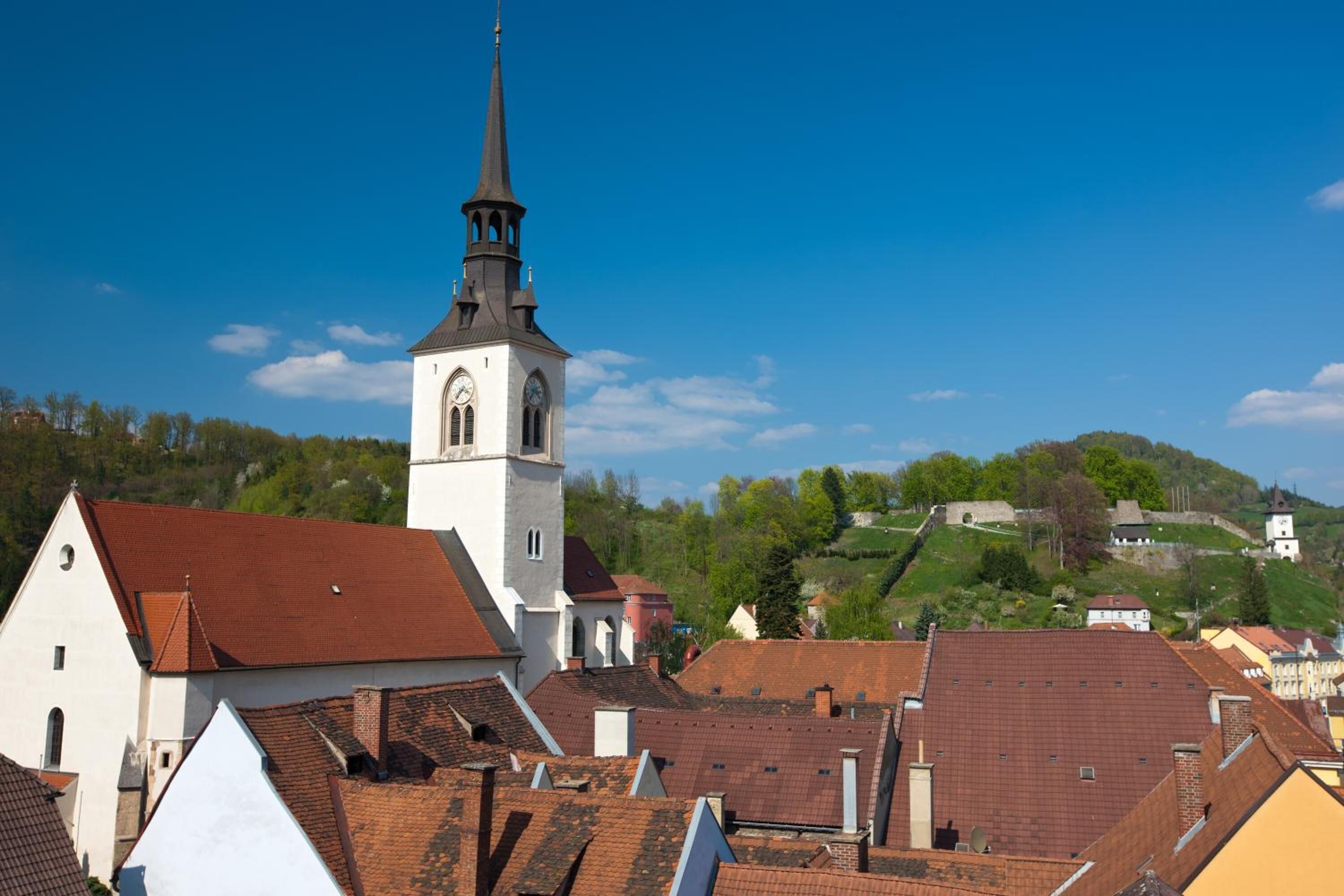 A church with a tall steeple and a clock tower, surrounded by buildings with red-tiled roofs.