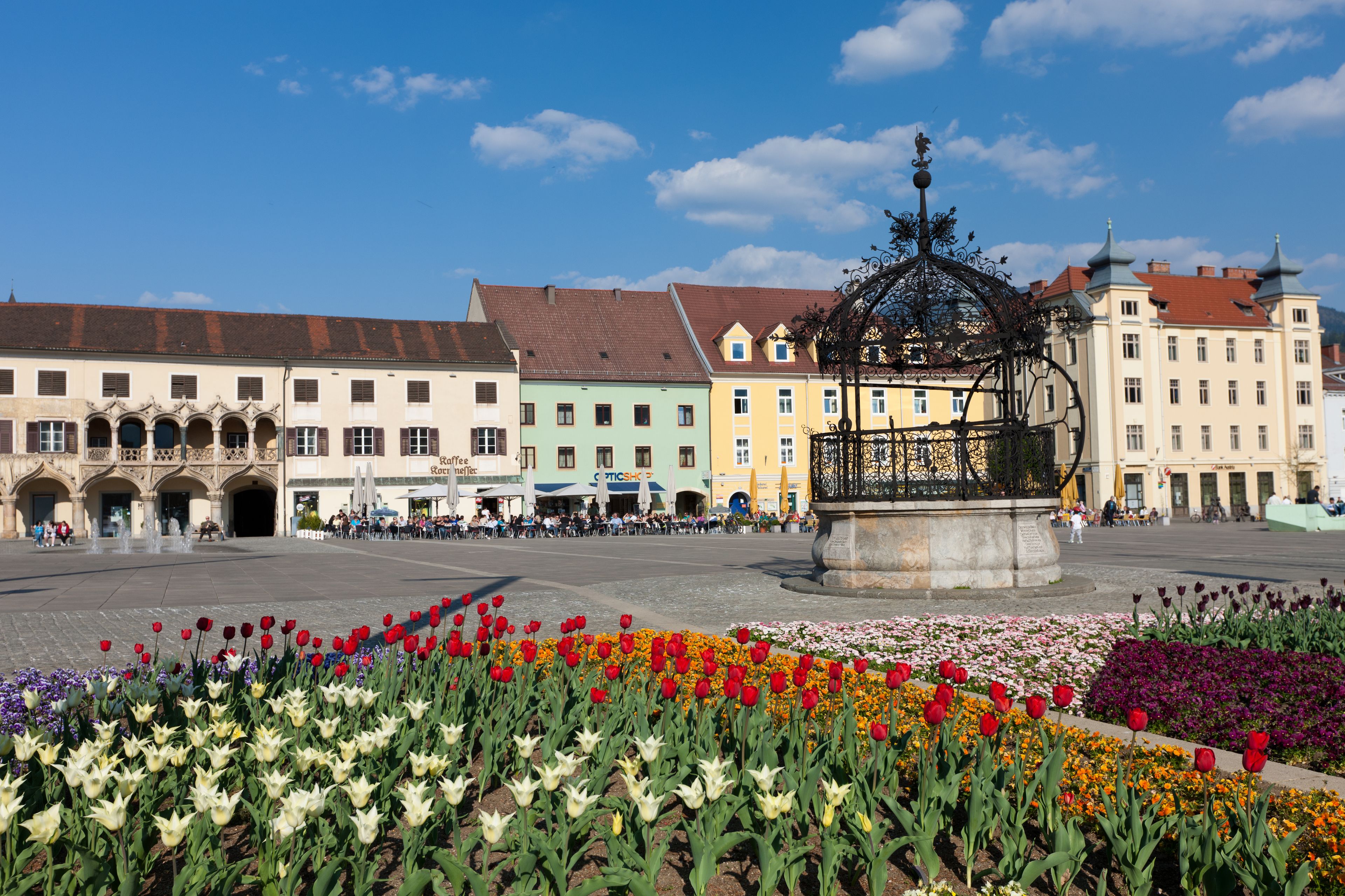A public square with a fountain, colorful buildings, and flower beds.