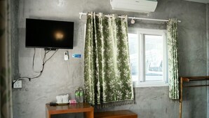 A room with a flat-screen TV mounted on the wall, a window with curtains, a wooden table with a telephone and a water bottle, and an air conditioning unit above the window. - PK Resort (Sikao)