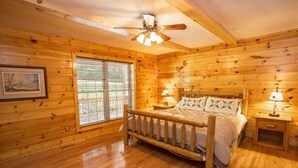 A wooden cabin bedroom with a bed, two bedside tables, a ceiling fan, and a large window with blinds. - Secluded Mountain Haven in Seven Devils (Seven Devils)