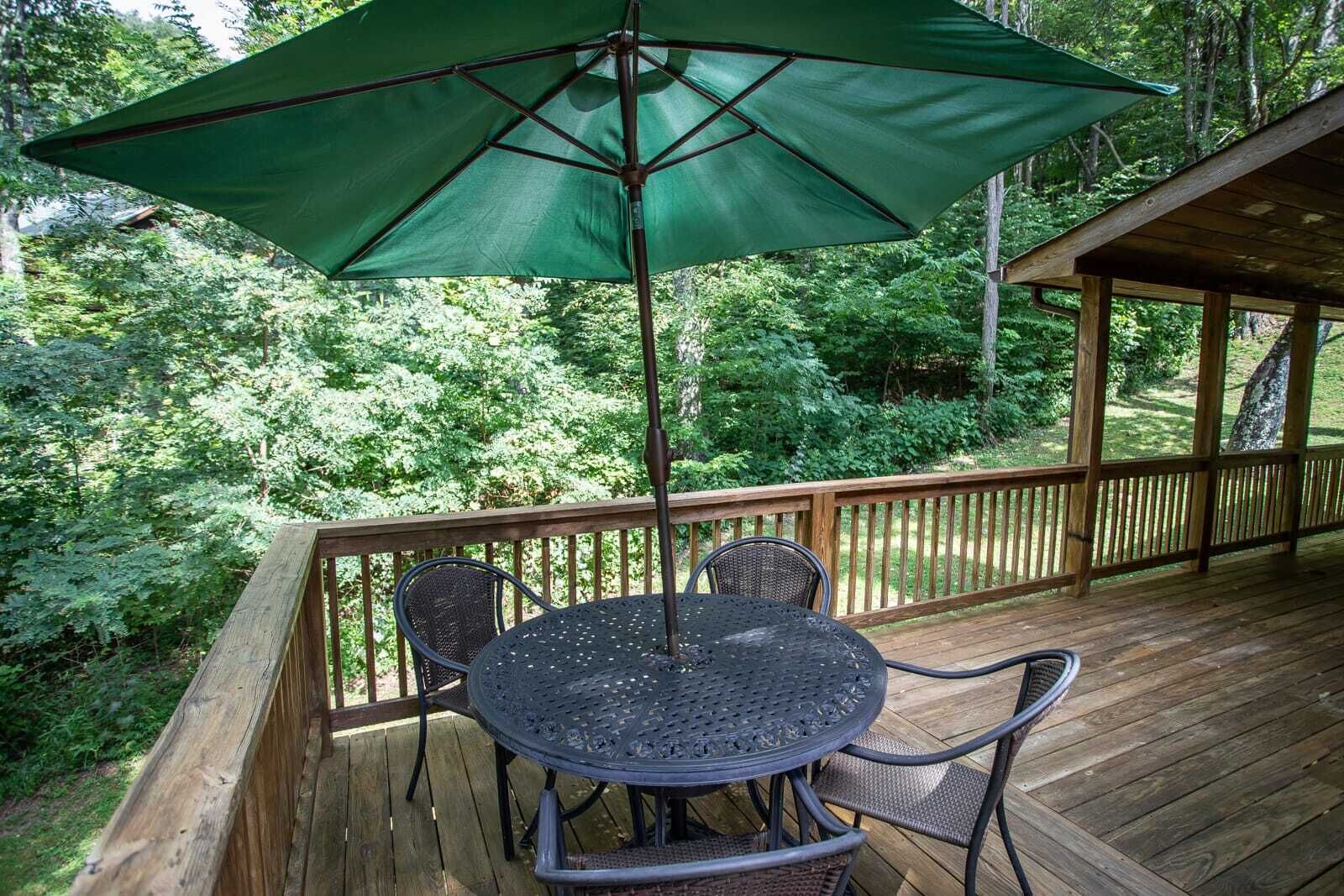 A wooden deck with a table and chairs, surrounded by a wooden railing, set in a lush green forest.