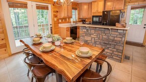 A wooden dining table set with plates and glasses, surrounded by chairs, in a kitchen with stone accents and wooden cabinets. - Secluded Mountain Haven in Seven Devils (Seven Devils)
