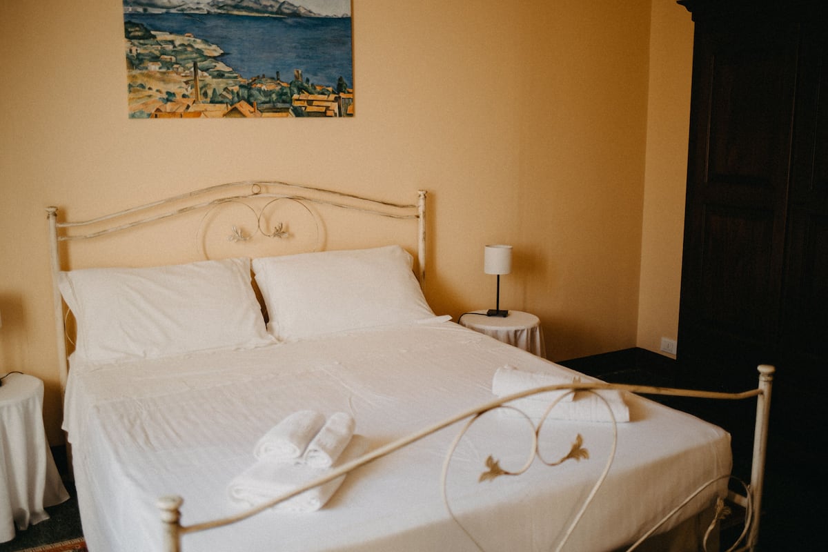 A neatly made bed with white linens and towels, a bedside table with a lamp, and a painting of a coastal scene on the wall.