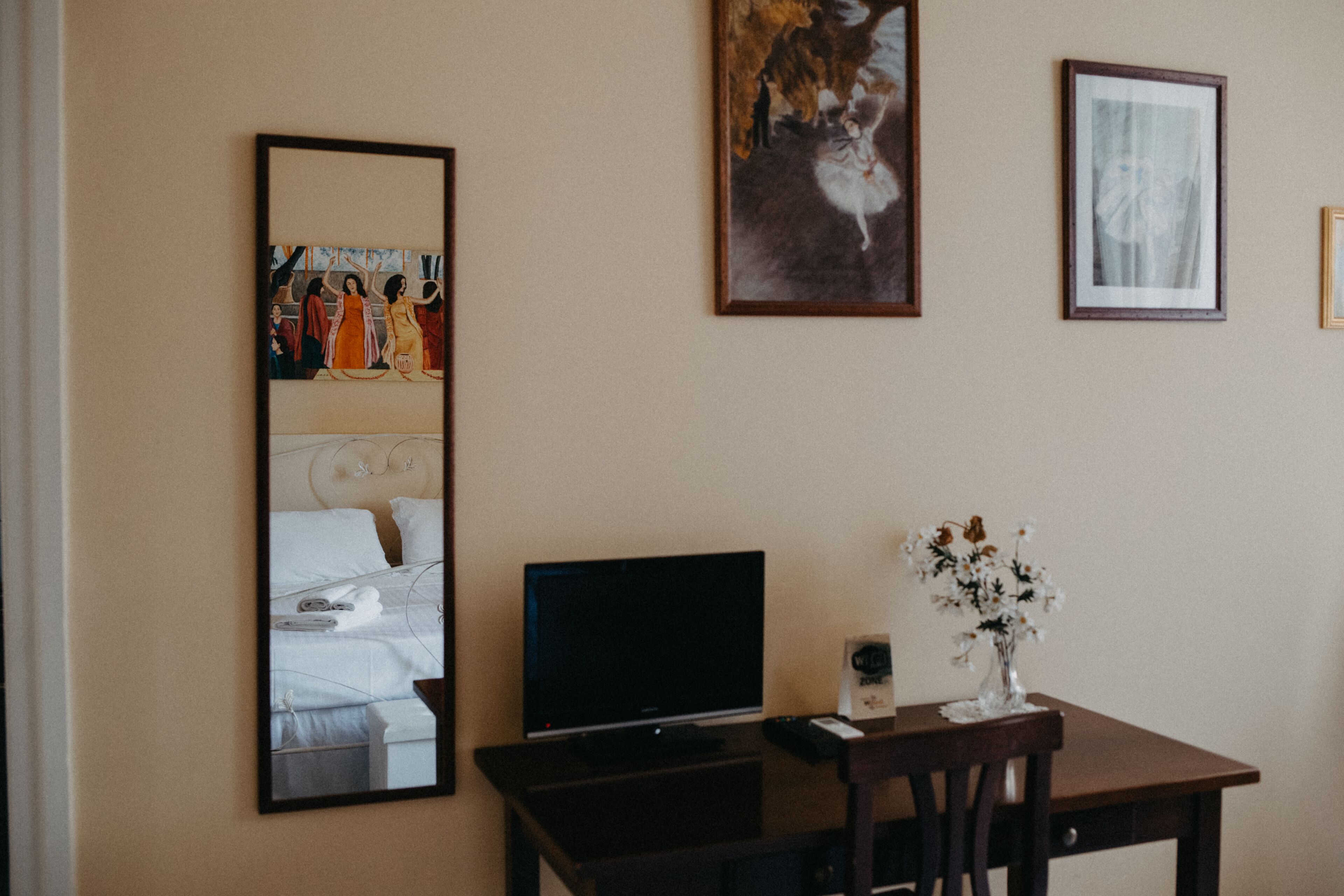 A hotel room with a bed, a TV, a desk with a vase of flowers, and framed artwork on the wall.
