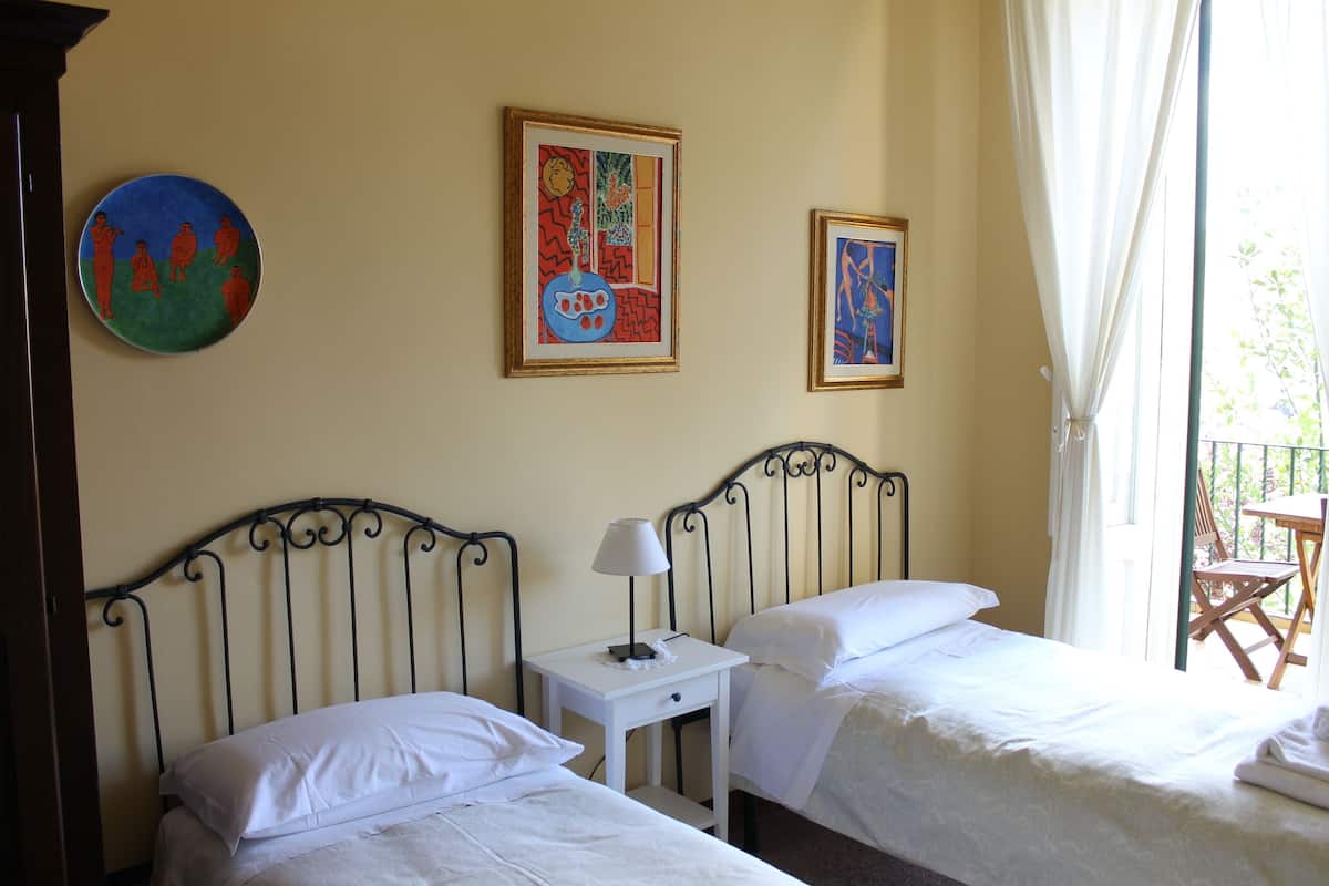 A hotel room with two beds, a small bedside table with a lamp, and two framed artworks on the wall.