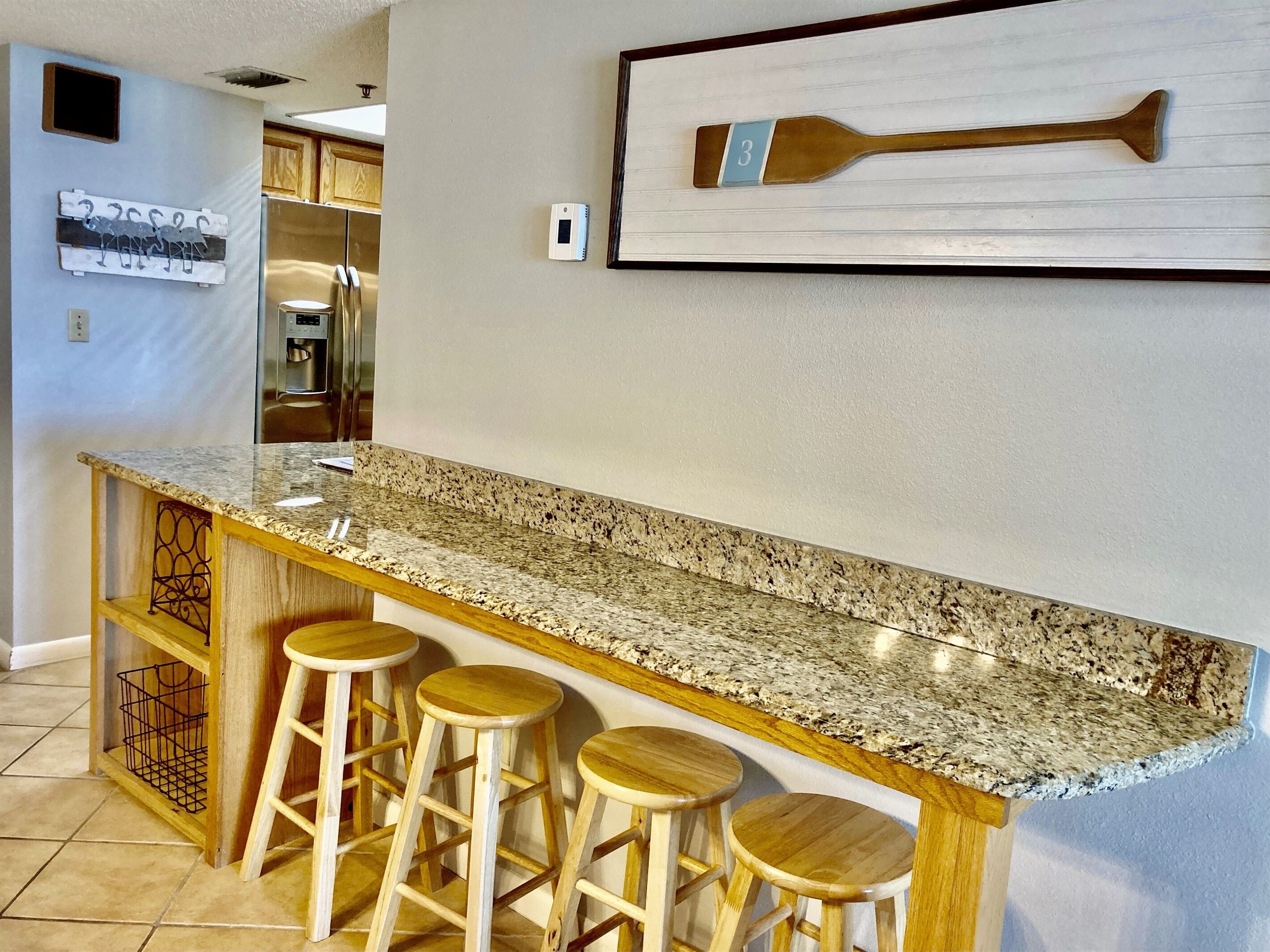 A kitchen with a granite countertop, wooden stools, a refrigerator, and a wall-mounted paddle.