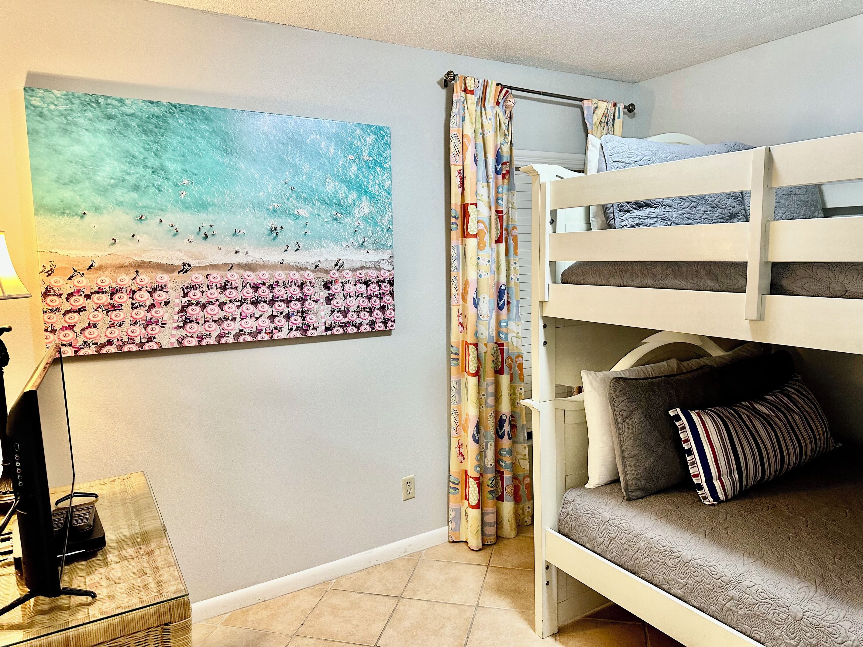 Une chambre avec un lit superposé, un tableau sur le thème de la plage, un rideau à motifs et une petite table avec une lampe.