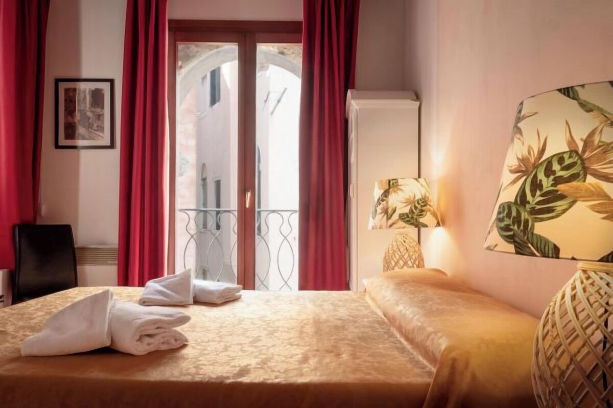 A hotel room with a bed, a chair, a balcony with red curtains, and a wall-mounted lamp.