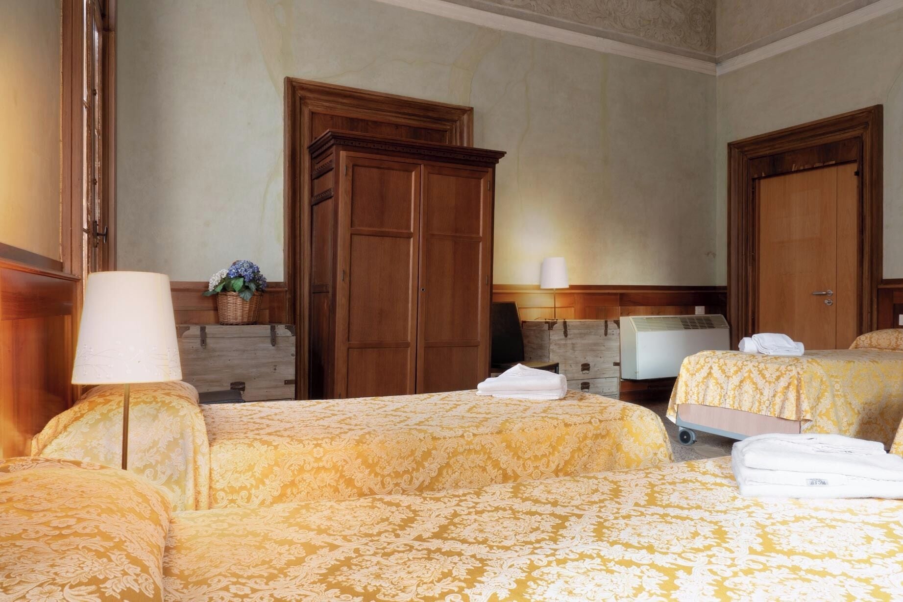 A hotel room with two beds, a wooden wardrobe, a lamp, a television, and a heater.