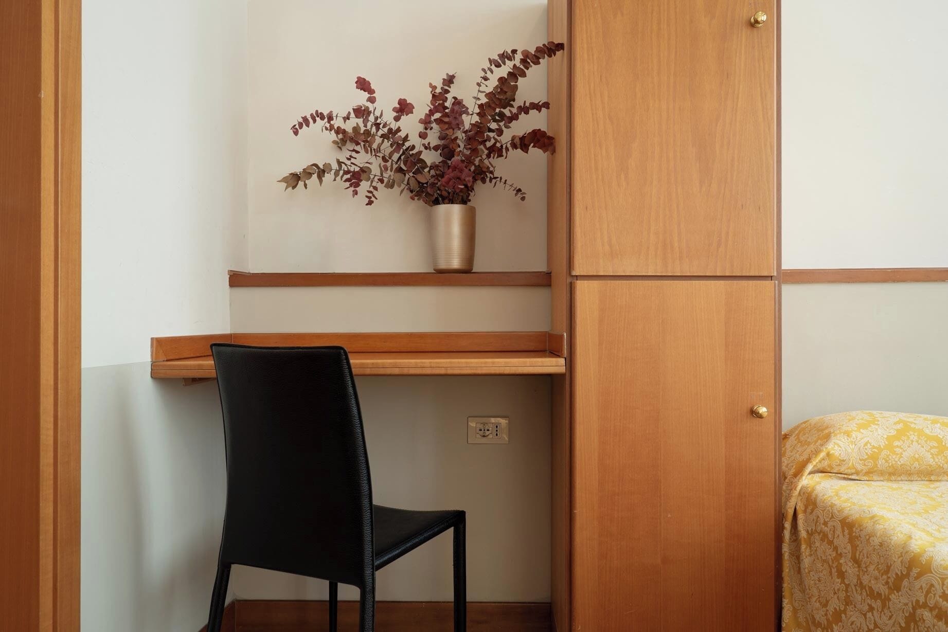 Una habitación con un escritorio de madera, una silla negra, una planta pequeña en un estante y una cama con cubrecolcha estampado.