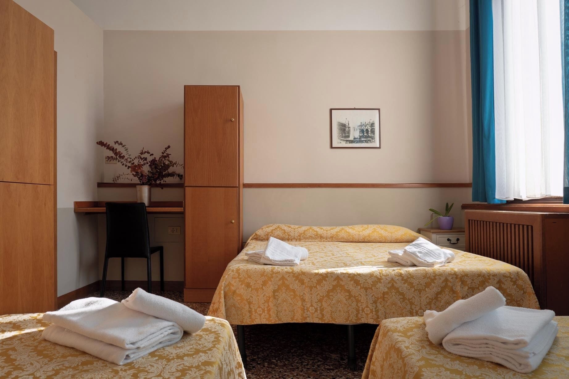 A hotel room with two beds, a desk, a chair, a wardrobe, a framed picture on the wall, and a window with curtains.