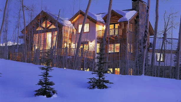 Une cabane en rondins dotée de nombreuses fenêtres et d’un balcon, entourée d’un paysage enneigé et d’arbres.