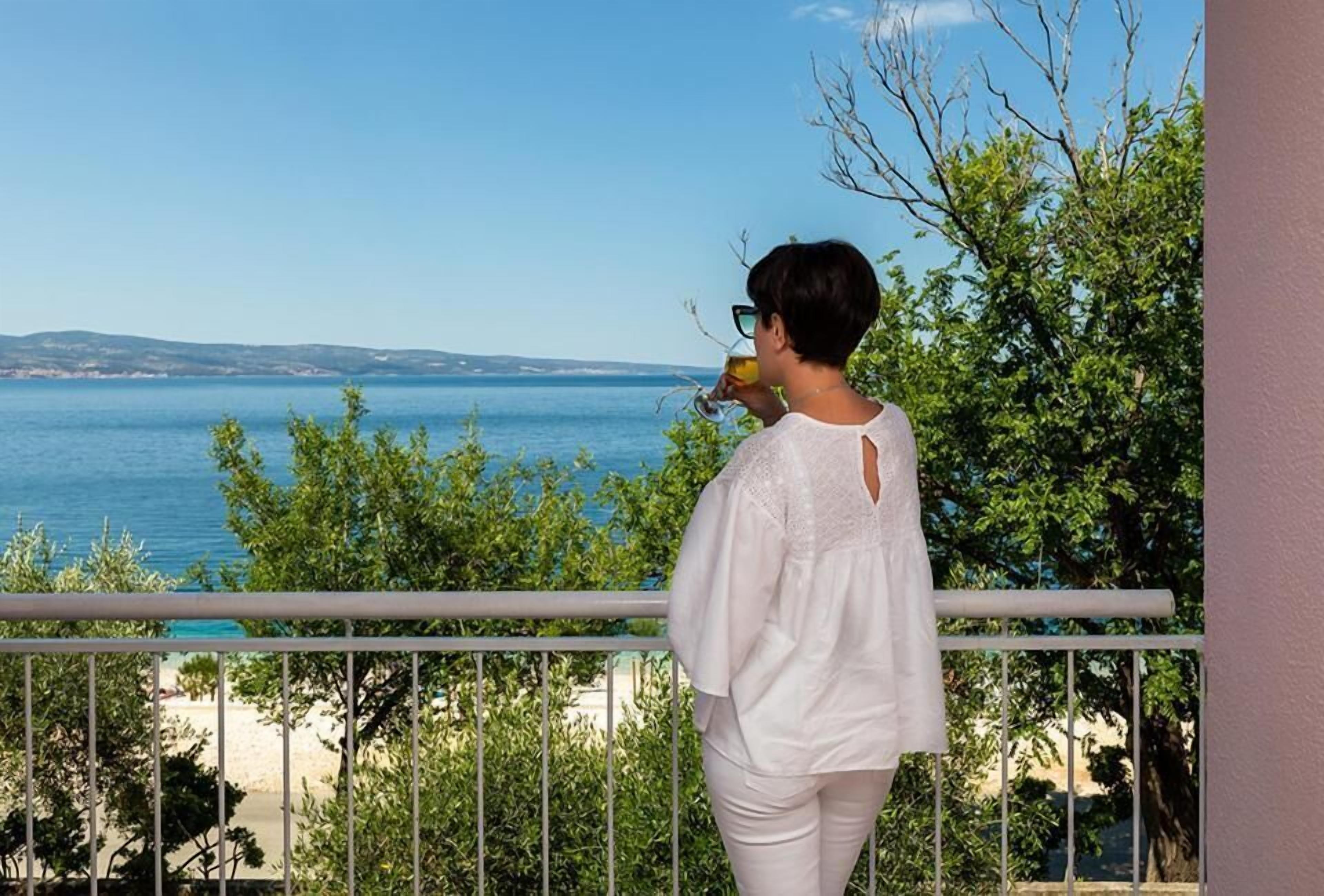 Une personne se tient sur un balcon qui donne sur une étendue d’eau, avec des arbres au premier plan.