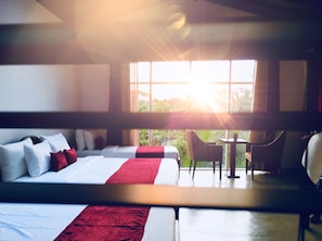 Habitación de hotel con una cama grande, una cama más pequeña, un escritorio y una silla. Se ve vegetación por la ventana.