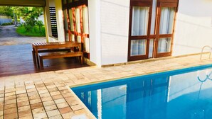 A swimming pool with a tiled deck, wooden bench, and a white building with large windows and a door. - 8 Plus  Villa (Battaramulla)