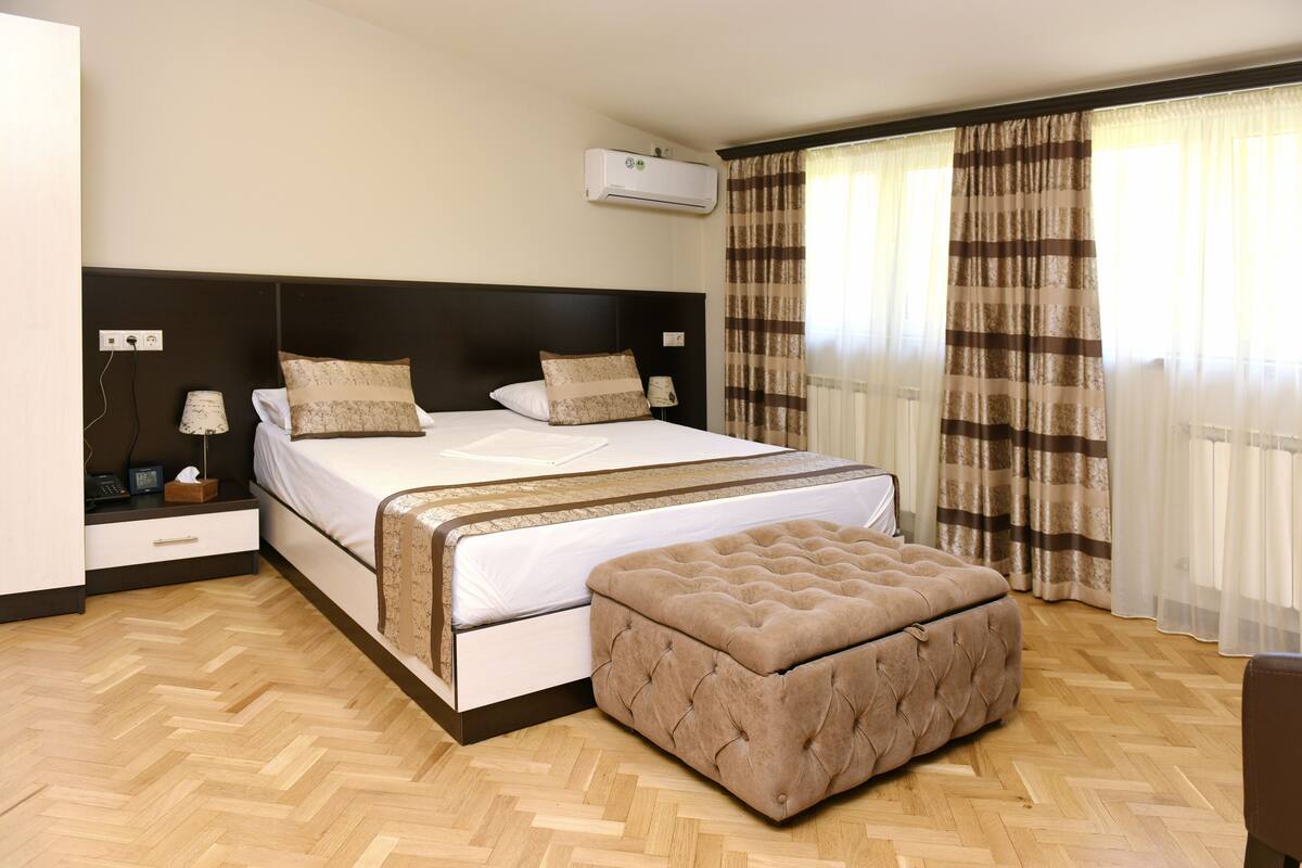 Une chambre d’hôtel avec un grand lit, un pouf, une table de chevet, une lampe et une fenêtre avec des rideaux.
