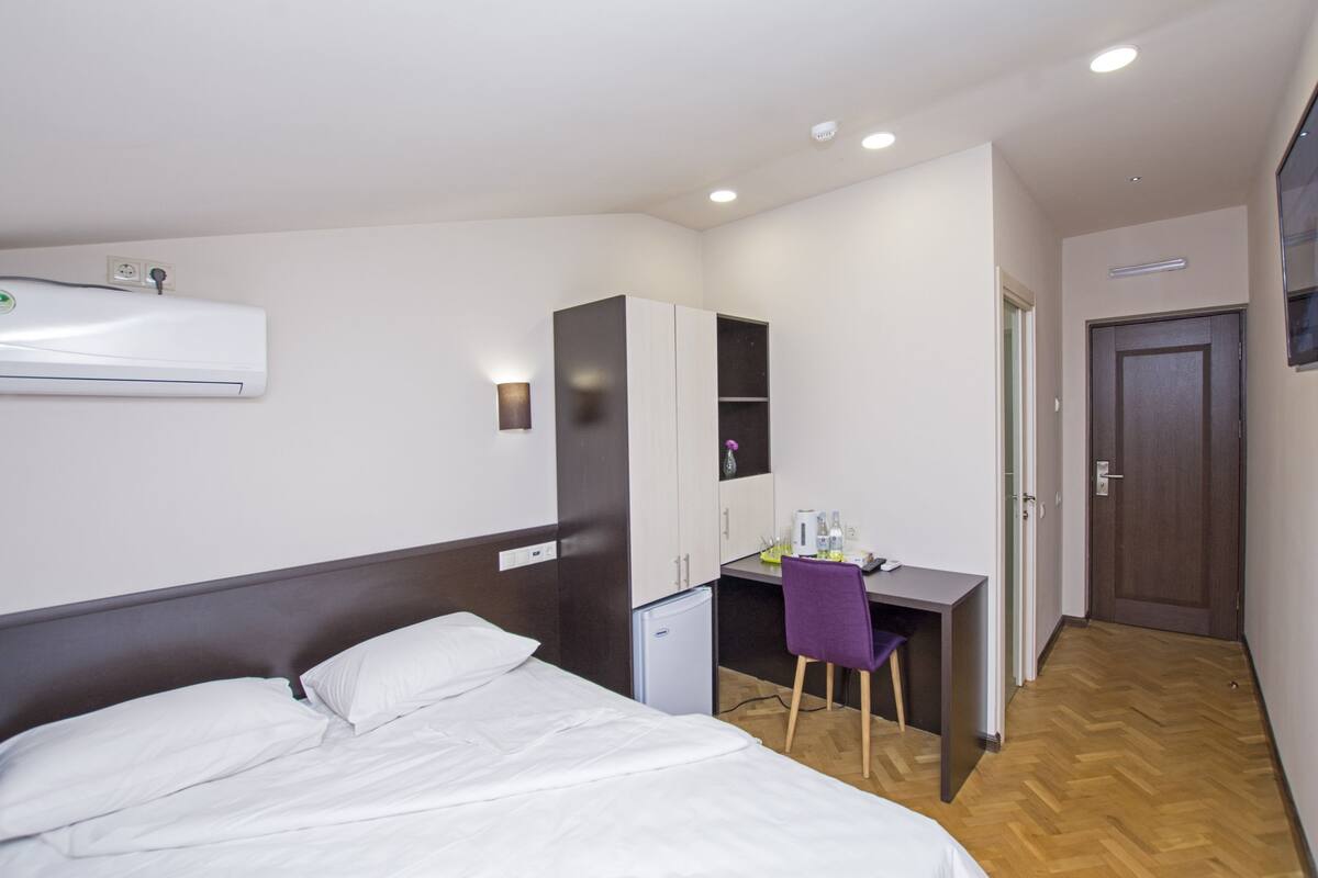 Une chambre d’hôtel équipée d’un lit, d’un bureau, d’une chaise, d’une armoire, d’un petit réfrigérateur et d’un appareil de climatisation.