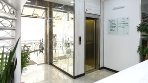 Um elevador moderno com painel de vidro, um aviso com números de quartos e comodidades, e um vaso de planta.