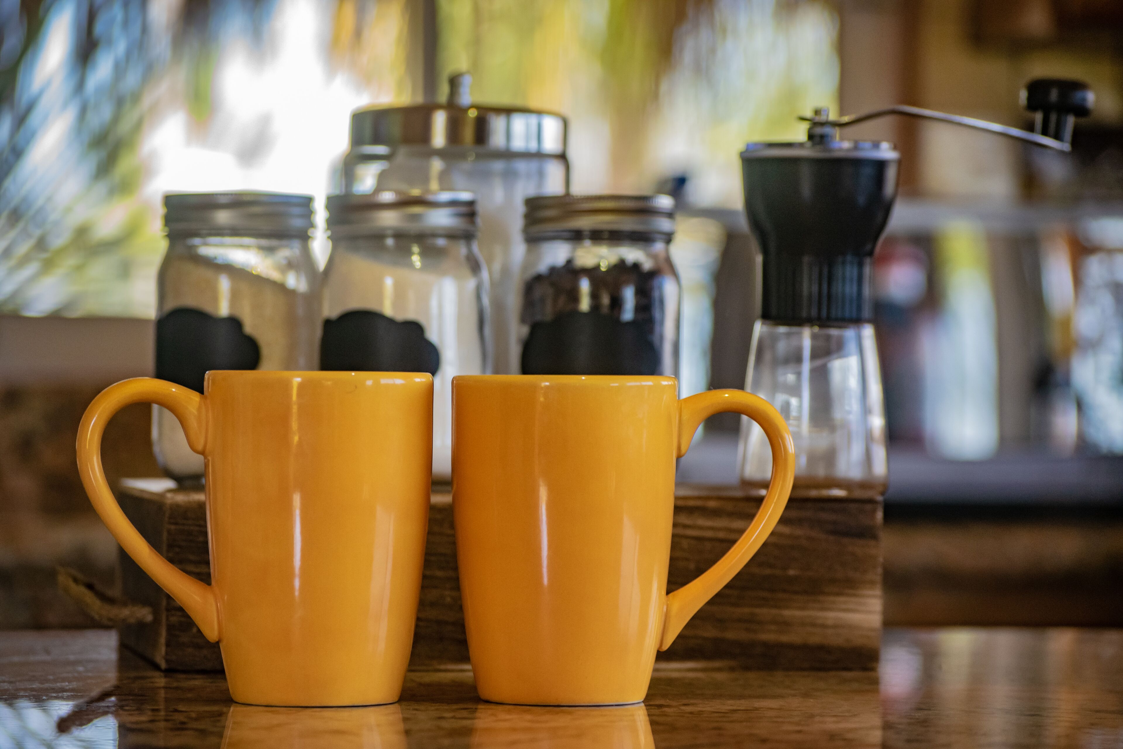 Deux tasses jaunes sur une surface en bois, avec des bocaux et un moulin à café en arrière-plan.