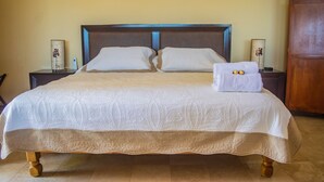 Una cama bien hecha con colcha blanca y falda marrón, flanqueada por dos mesitas de noche con l ámparas, en una habitación con cabecera de madera y una puerta de madera.