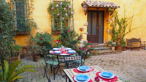 An outdoor patio with a table set for two, surrounded by potted plants and a yellow building with a tiled roof. - Two apartments for groups or families up to 9 In St. Peter area, courtyard (Roma)