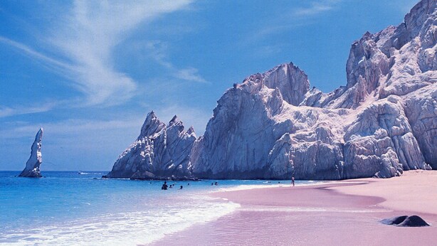 Une plage au rivage de sable rose, avec une eau d’un bleu cristallin et des falaises rocheuses en arrière-plan.