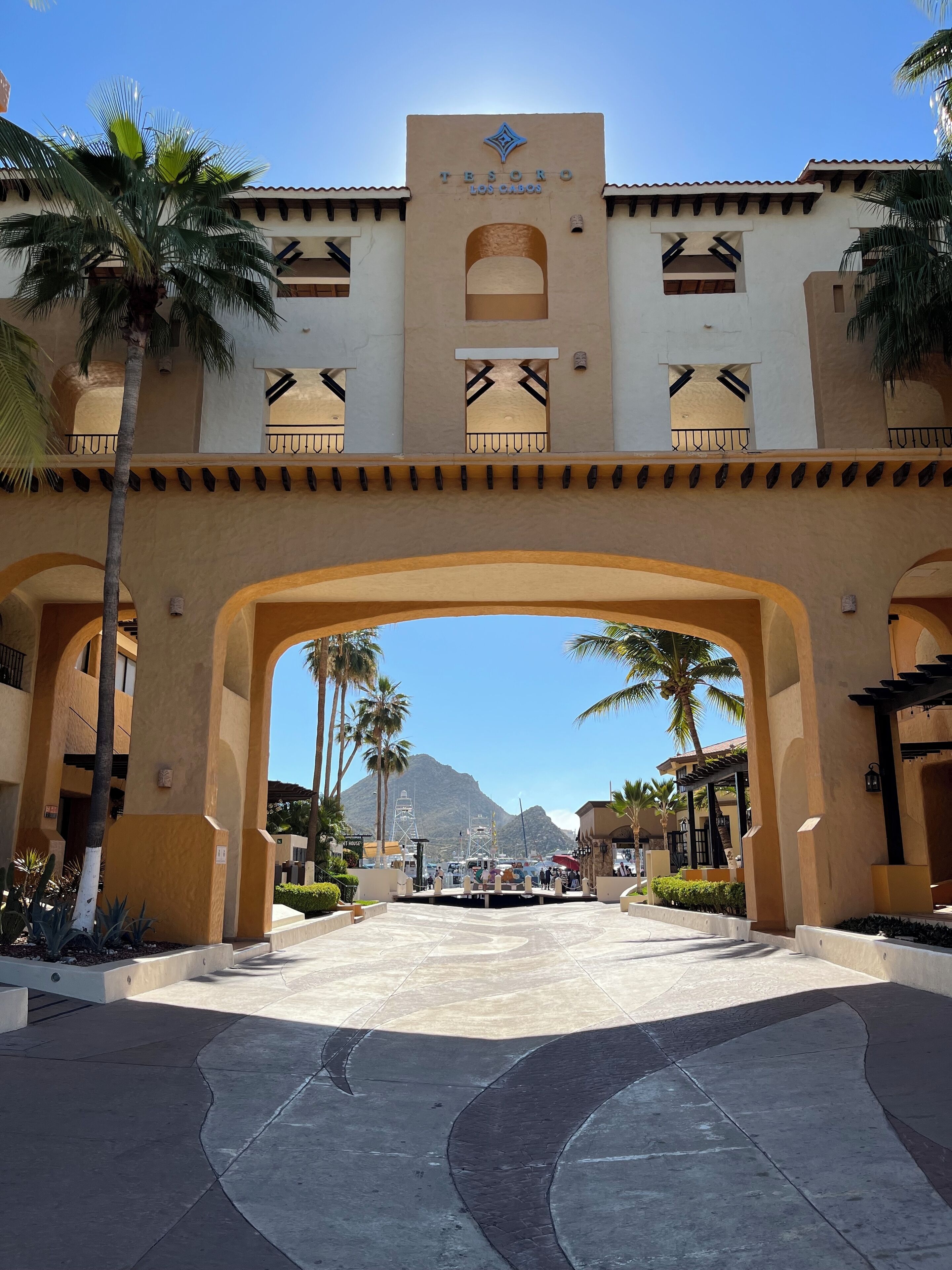 Entrada de un hotel con un gran arco, palmeras y montañas al fondo.