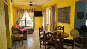 Une pièce aux murs jaunes, avec un ventilateur au plafond, un lit recouvert d’une couverture rouge, une table avec des chaises et une fenêtre avec des rideaux.