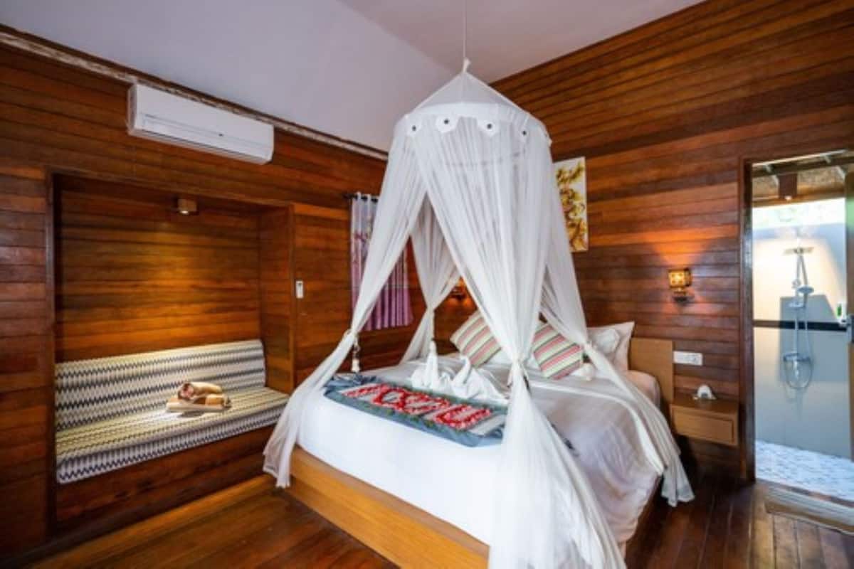 Ein Zimmer mit Holzwänden, einem Bett unter einem weißen Baldachin, einer Bank mit Decke und einem Nachttisch aus Holz mit einer Lampe.