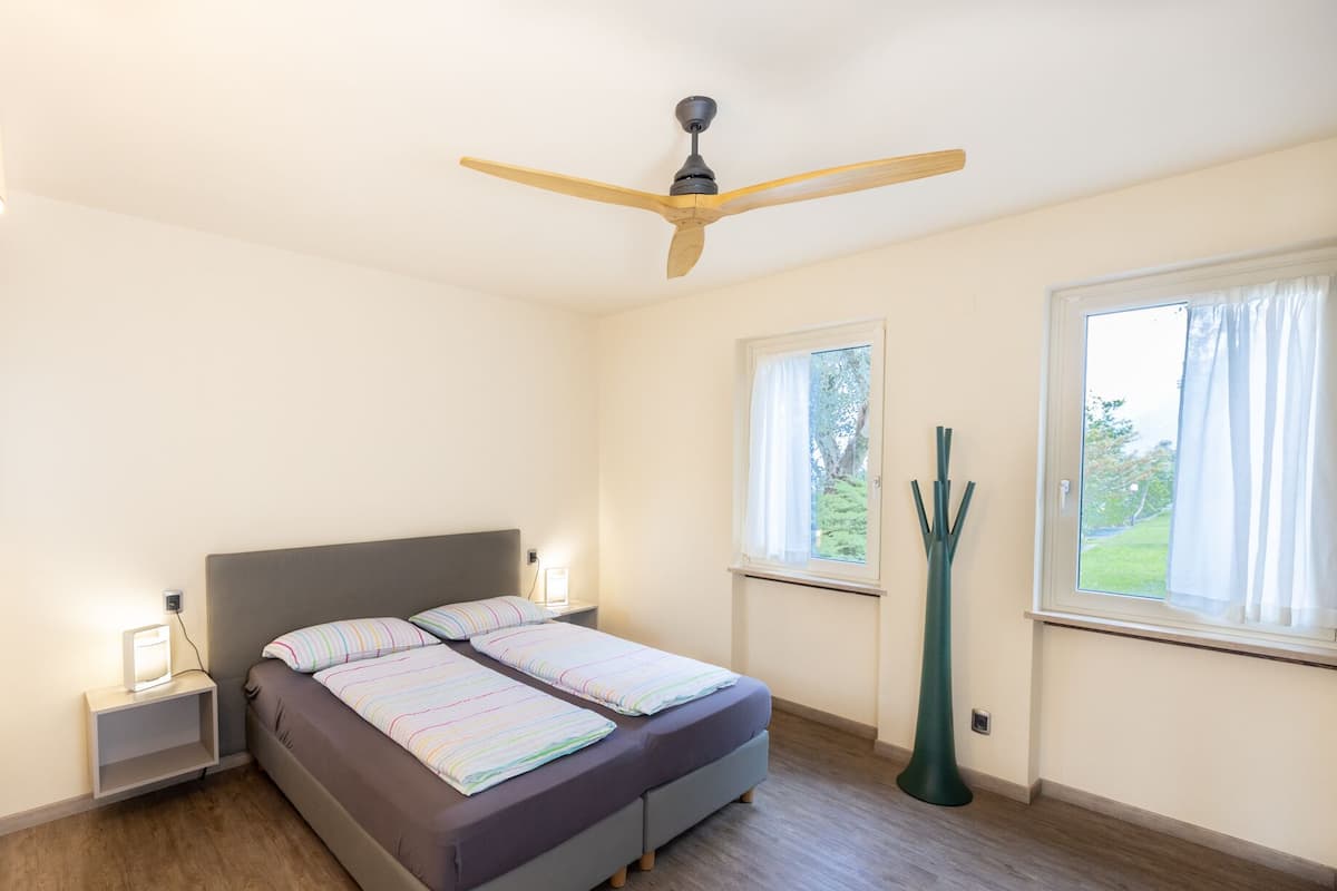 A bedroom with a bed, a ceiling fan, bedside lamps, and windows.