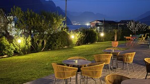 An outdoor patio area with wicker chairs and tables, a well-maintained lawn, and a view of mountains in the background. - Residence Alesi (Malcesine)
