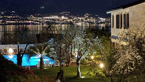 A nighttime view of a waterfront property with a pool, trees, and a building with columns. - Residence Alesi (Malcesine)