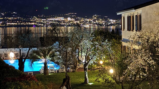 A nighttime view of a waterfront property with a pool, trees, and a building with columns. - Residence Alesi (Malcesine)