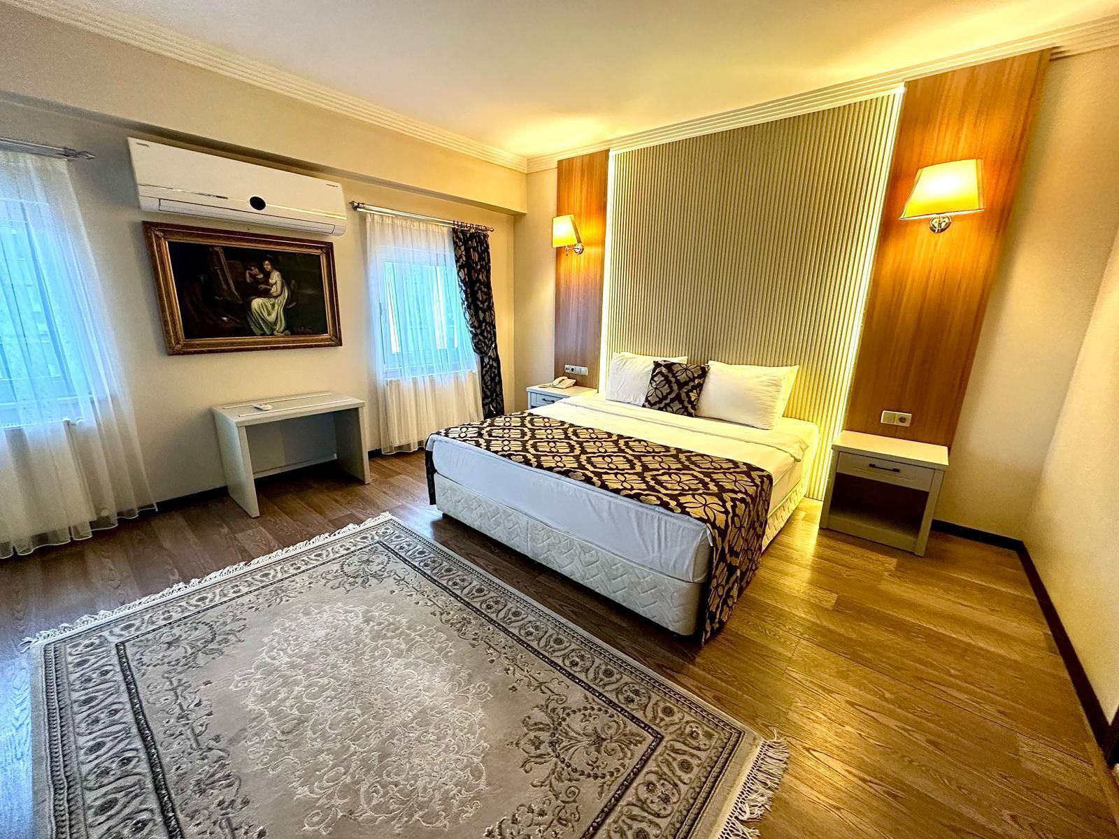 Una habitación de hotel con una cama, mesitas de noche, un escritorio, un cuadro y una alfombra estampada.