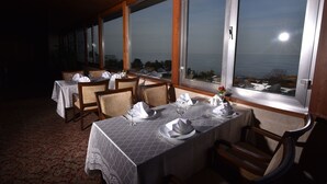 A dining area with tables set for two, large windows with a view of the sea, and a wooden ceiling. - Saylamlar Hotel (Trabzon)