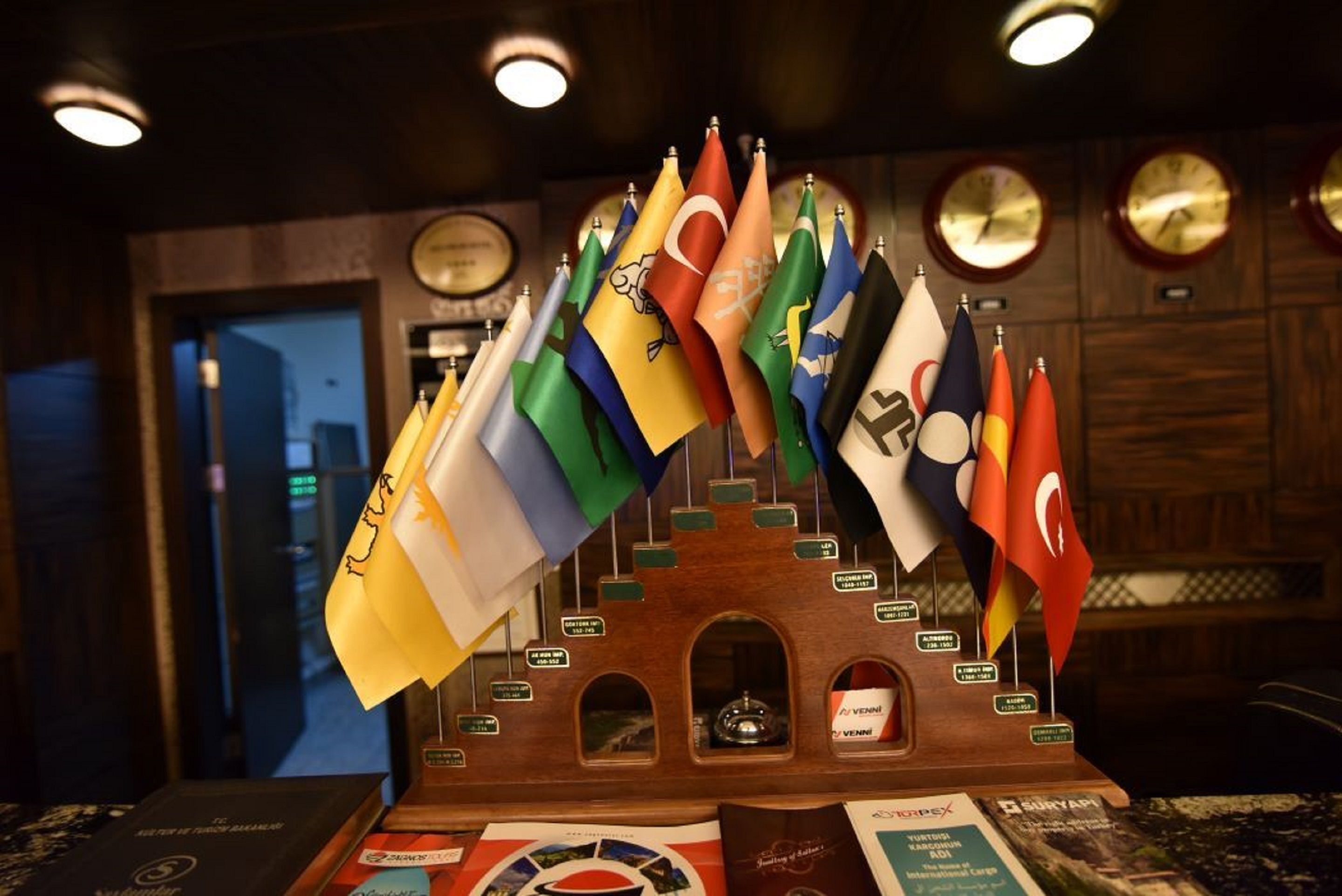 Una exhibición de varias banderas nacionales colgadas de una estructura de madera con placas, en una sala con relojes en la pared.