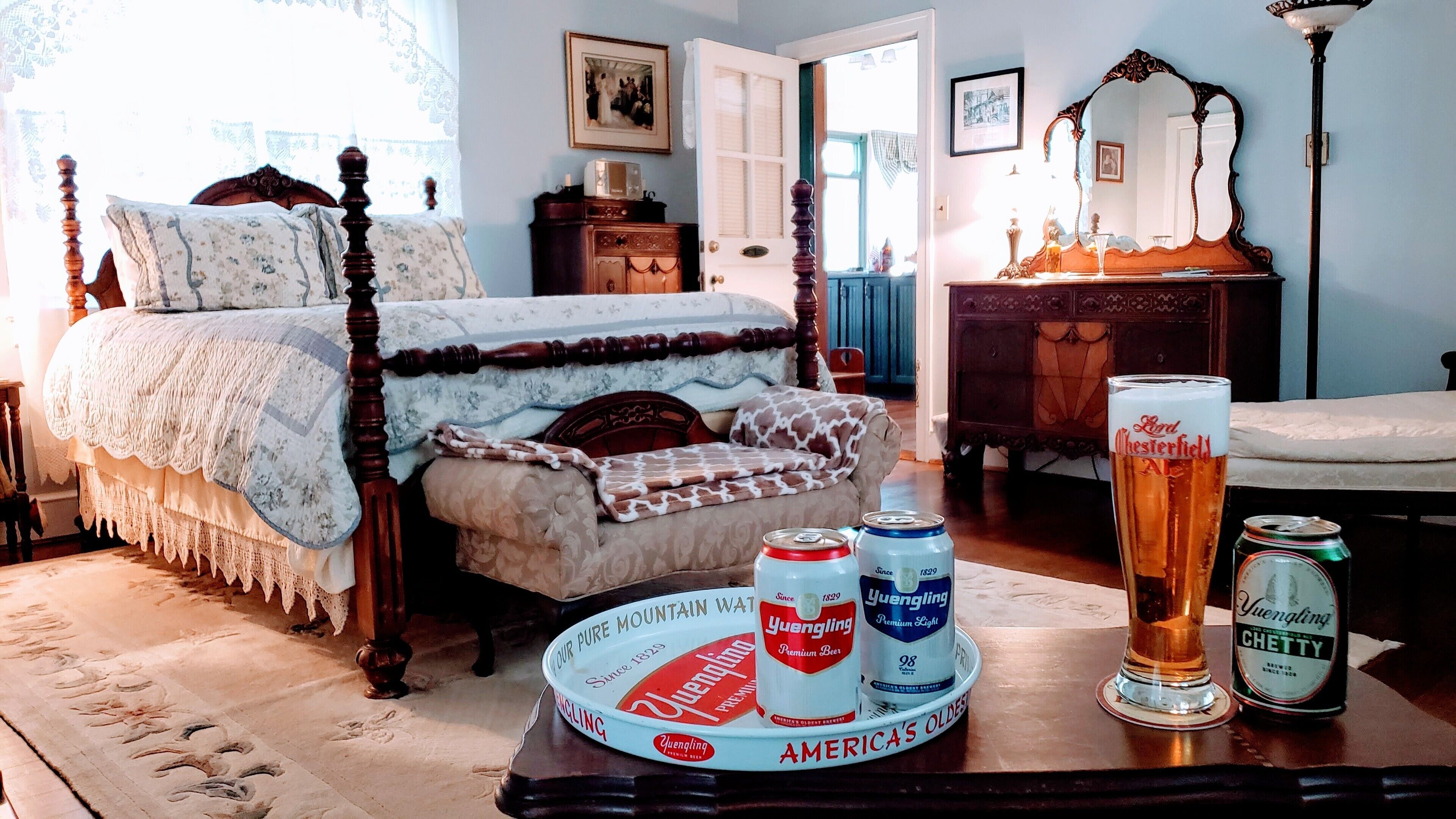 Une chambre à coucher avec un lit à baldaquin, une commode en bois, un miroir et une table sur laquelle se trouvent des canettes de bière Yuengling et un verre.