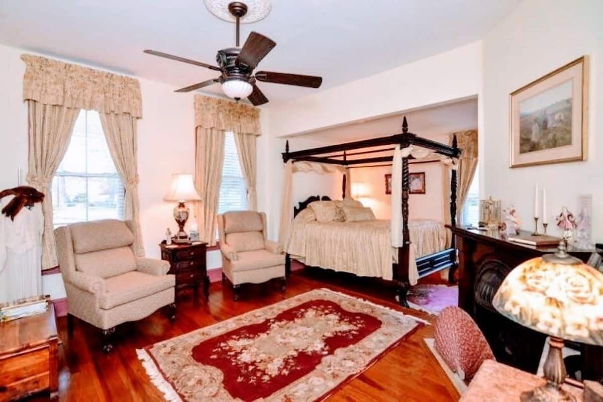 A bedroom with a four-poster bed, two armchairs, a wooden floor, and a ceiling fan.