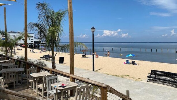 Vista frente al mar con una terraza de madera, mesas y sillas, que da a una playa de arena y un mar tranquilo.